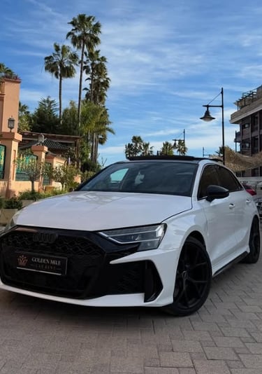 White Audi RS3 luxury hatchback parked on a paved street with palm trees and Mediterranean architecture in the background.