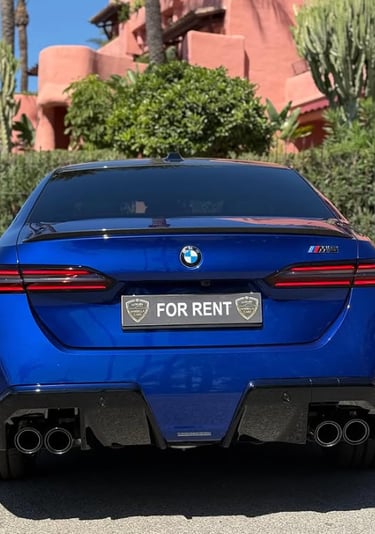 Rear view of a blue BMW M5 luxury rental car parked in front of a Mediterranean villa.