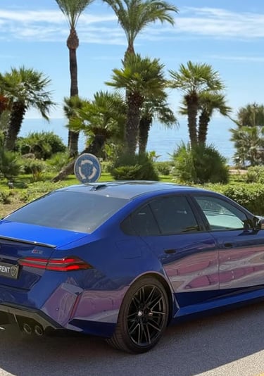 A blue BMW M5 luxury sedan for rent parked near tropical palm trees and a scenic ocean coastline.