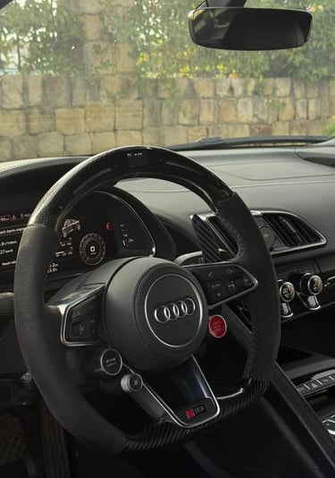 Interior view of an Audi R8 luxury sports car showing the carbon fiber steering wheel and digital dashboard.
