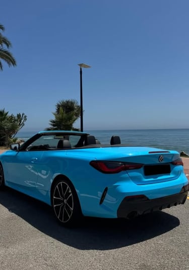 A bright blue convertible BMW parked on a coastal road with palm trees and a view of the ocean.