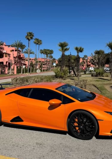 Orange Lamborghini Huracan supercar with black wheels parked near tropical palm trees and pink buildings.