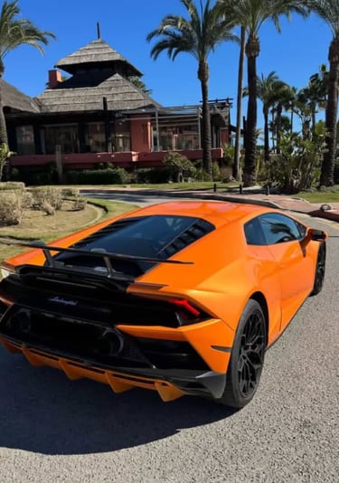 Orange Lamborghini Huracan supercar parked on a sunny road near a tropical resort with palm trees.