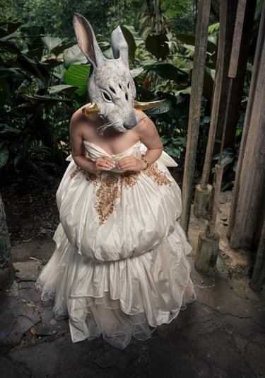 A person wearing a mystical white animal mask and elegant cream ballgown in a lush jungle setting.