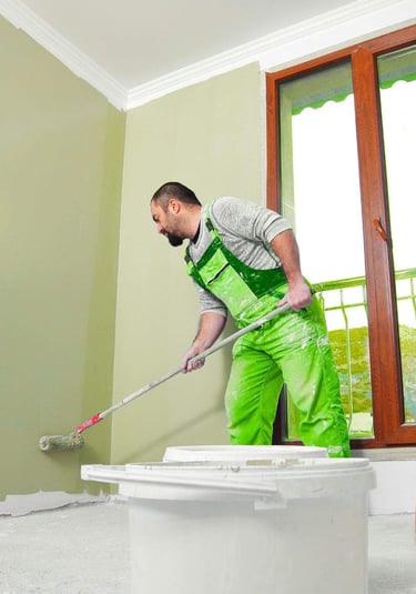 Person painting an interior wall with a roller near a window.