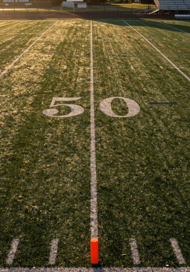 Wide environmental shot looking down the length of a youth flag football field at golden hour, yard lines painted on grass, portable orange pylons at the end zone corners, empty bleachers visible at right edge, long shadow lines stretching across the turf from low sun at left frame