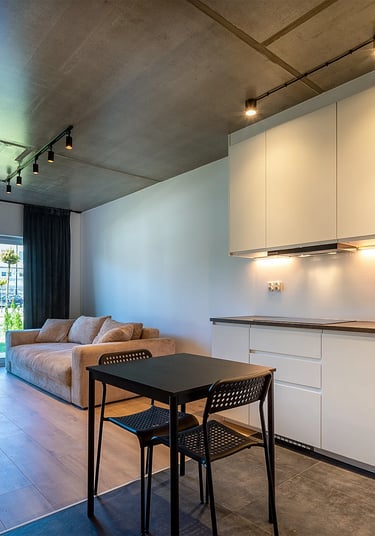 Nowoczesny salon z aneksem kuchennym: białe szafki, stół z krzesłami, sofa, drzwi balkonowe.