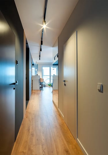 Korytarz z drzwiami po obu stronach, drewniana podłoga, listwowe oświetlenie sufitowe okno na końcu.