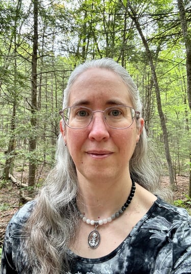Portrait of a middle-aged white woman with long gray hair and glasses, standing in the woods.
