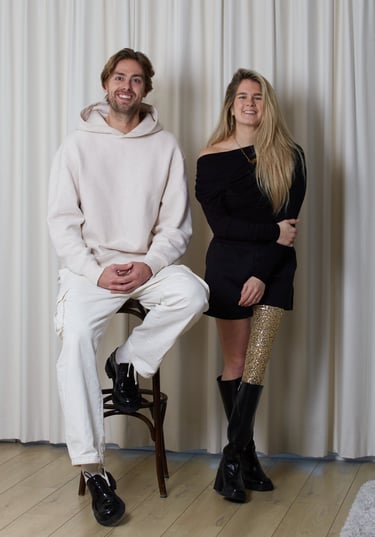 Smiling man in a white hoodie and woman with a gold prosthetic leg posing against white curtains.