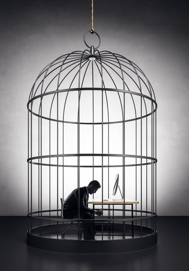 a man hunched over at a desk that is inside a large birdcage it is in grayscale 