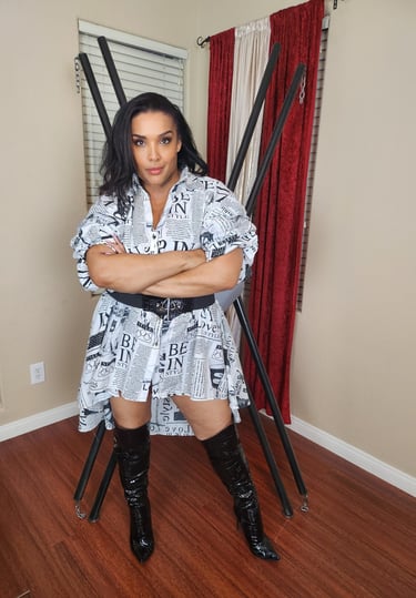 MsMadyson in a newsprint dress and over the knee boots is standing in front of a St. Andrews cross