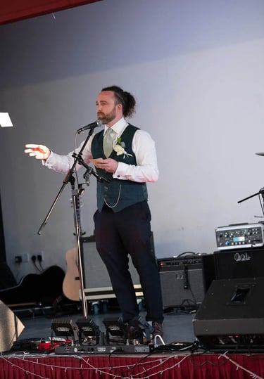 A best man in a green waistcoat giving a wedding speech into a microphone on stage.
