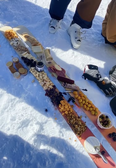 A long charcuterie board spread laid out across a pair of skis on a snowy mountain, featuring variou