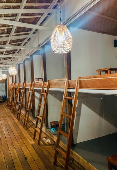 Rustic wooden capsule dorm beds with ladders and wicker pendant lighting in a cozy tropical hostel.