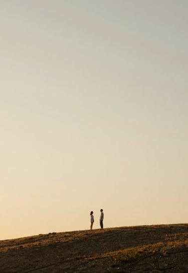 A wide sun-drenched shot of a couple standing on a hill at golden hour, cinematic wide angle, soft #F7F2EB sky tones and warm shadows.