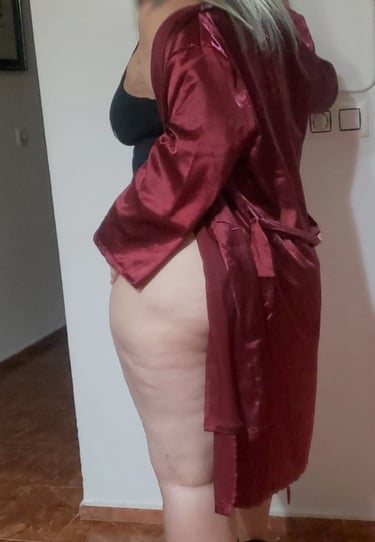 Escort madura colombiana en bata roja de satén, de rodillas sobre el sofá, mostrando sus piernas provocativas.