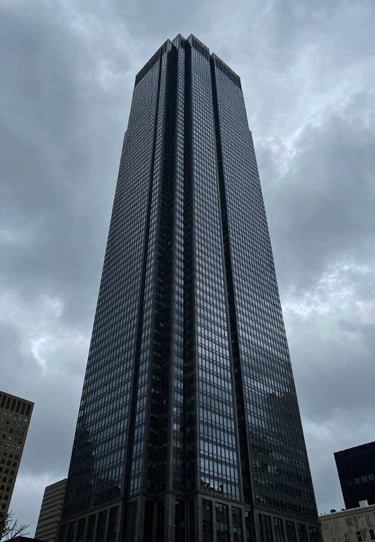 Low angle photography of a sleek, modern skyscraper in a US metropolis under a heavy, cinematic overcast sky. The style is professional and visually engaging with deep charcoal and blue tones.