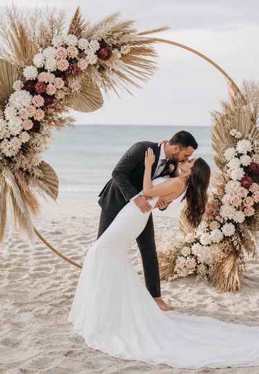 A bride and groom kiss under a floral beach wedding arch with pampas grass and roses.