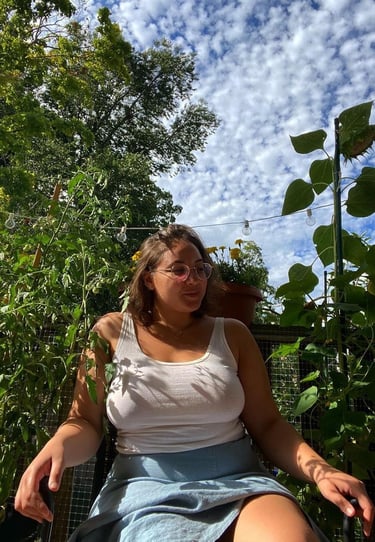 A woman wearing glasses and a tank top sits in a lush garden under a blue sky with clouds.