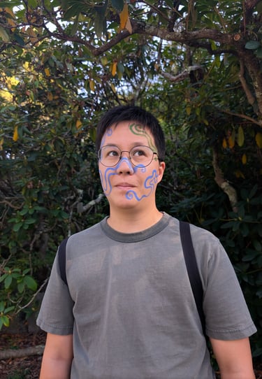 Person with blue and green artistic face paint patterns posing outdoors in front of leafy trees.