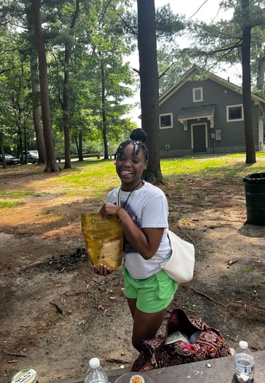 A smiling young woman holding a snack bag at an outdoor park with a cabin and tall pine trees.