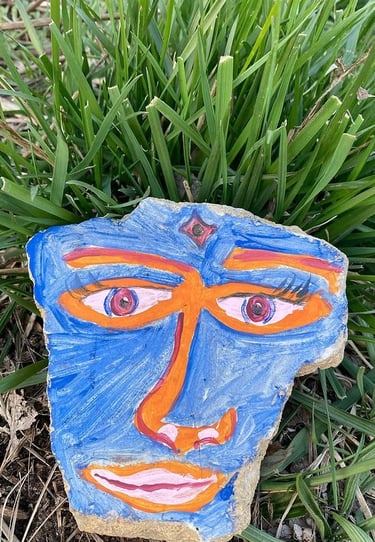 Hand-painted rock art featuring a blue face with orange accents nestled in green grass.