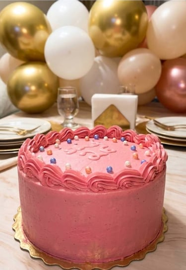 pastel de cumpleaños color rosa y de fondo una decoración de globos en dorado.