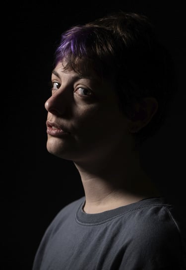 Dramatic low-key portrait of a person with short hair and purple highlights against a black background.