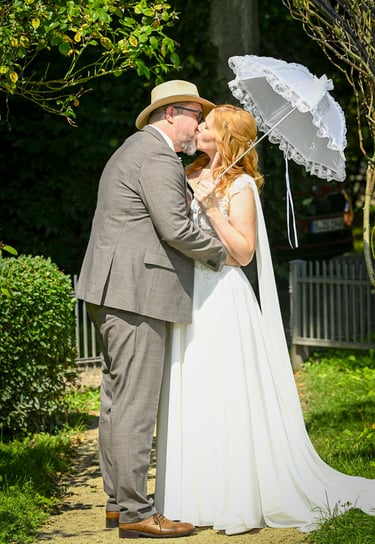 Küssendes Brautpaar im Grünen mit weißem Spitzenschirm nach der Hochzeit.