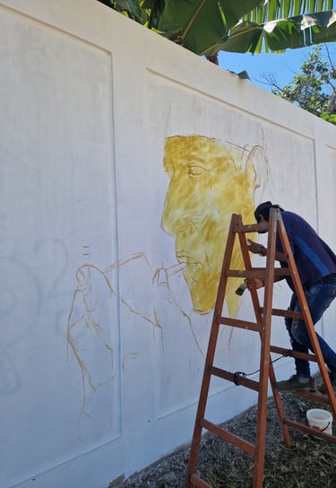 Artist painting a large yellow mural portrait on a white outdoor wall at Mokanranko Recreo
