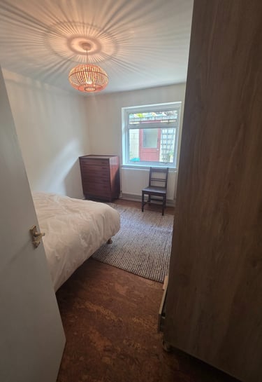 A bright bedroom with a woven pendant light, wooden dresser, and window view of a patio.