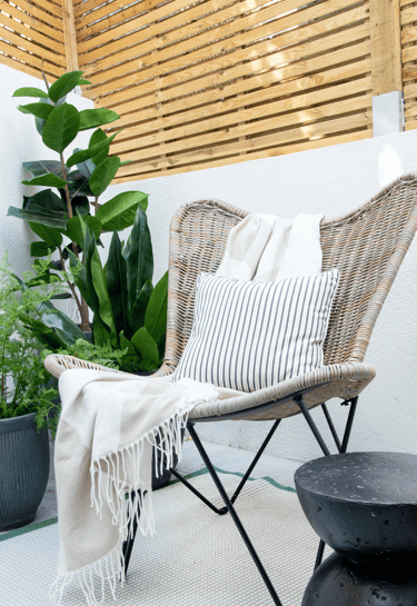 Wicker outdoor lounge chair with striped pillow and throw blanket on a modern patio with green plants.