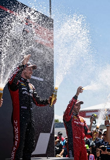 A low-angle photography shot of a motorsport podium celebration. Bright white champagne spray fills the air, catching the sunlight, with hints of red team gear visible in the crowd, North American / US racing event.