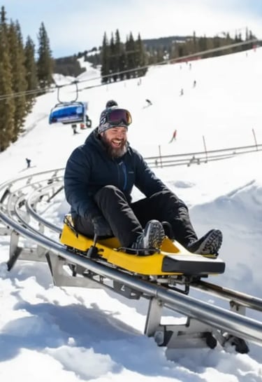 A man in a dark winter jacket smiling while riding a yellow mountain coaster down a snowy slope with