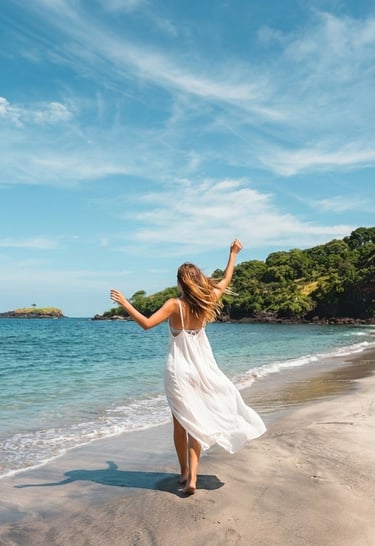 A woman in a flowing white dress stands barefoot on the sandy shore of Virgin Beach, Karangasem, Bali, facing the turquoise s