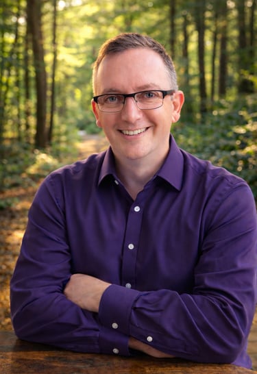 Smiling man with glasses in a purple shirt posing for a professional outdoor headshot in a forest.