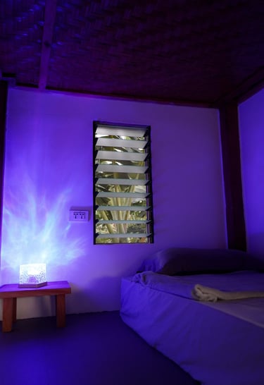 A cozy bedroom illuminated by a purple ambient lamp next to a jalousie window.