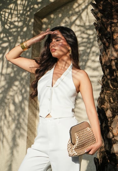 A stylish woman in a white halter vest and trousers poses with a gold woven clutch bag.