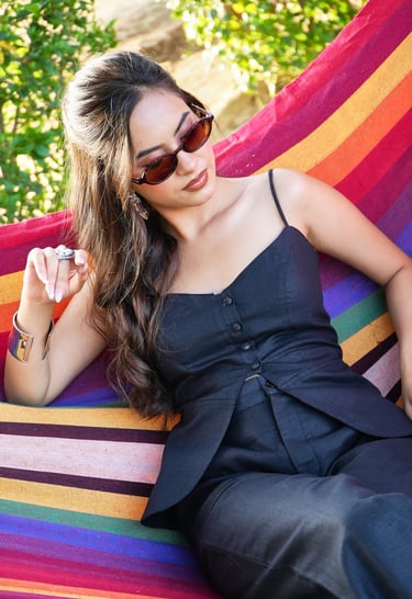 A stylish woman in a black linen outfit and sunglasses relaxing on a colorful striped hammock outdoors.