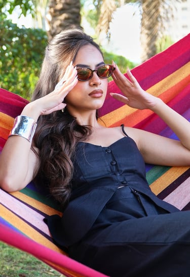 A fashionable woman in a black outfit and sunglasses relaxes in a colorful striped hammock outdoors.
