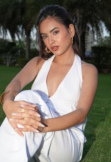 A woman posing in a white halter top and gold jewelry on a lush green lawn.