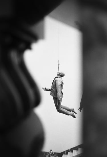 Black and white artistic photograph of a surreal human sculpture hanging by a rope in a gallery.