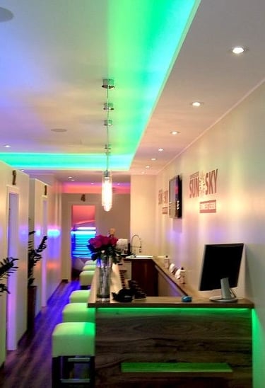 Zabudowa w salonie opalania z kolorowym podświetleniem LED, drewnianym blatem recepcji i nowoczesnym sufitem z punktowym oświ