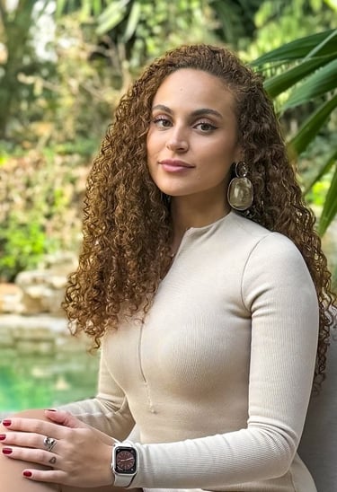 Dalli, founder of The Studio D, sitting outdoors in a cream top with curly hair and statement earrin