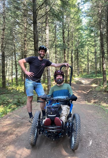 Sean seated in his 3-wheeled adaptive mountain bike on a forest trail with his friend Alex standing