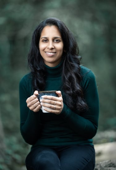 Shivani gently smiling, holding a mug and sitting in a woodland