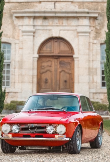 An Image of a red 1974 Alfa Romeo GTV parked between to rows of trees in an elegant setting.