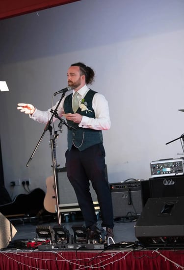 A best man in a green waistcoat giving a wedding speech into a microphone on stage.