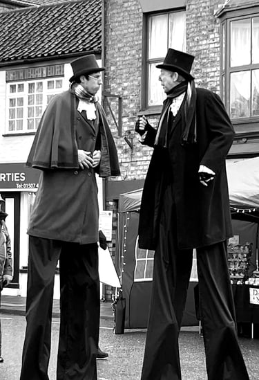 Two street performers on high stilts wearing Victorian costumes and top hats at a festival.